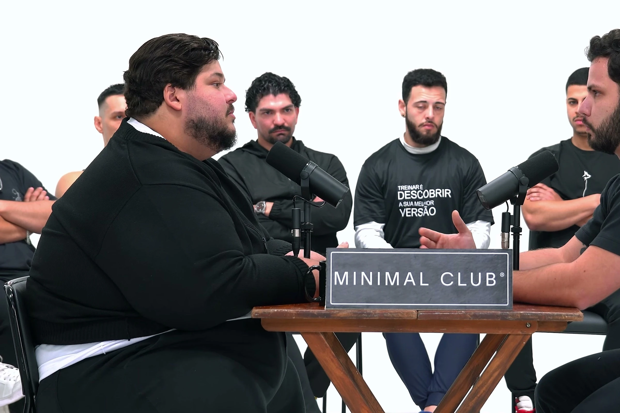 A mesa de pinho do 1 vs 30 com a placa da Minimal Club: o convidado e um dos trinta frente a frente, com a roda atrás.