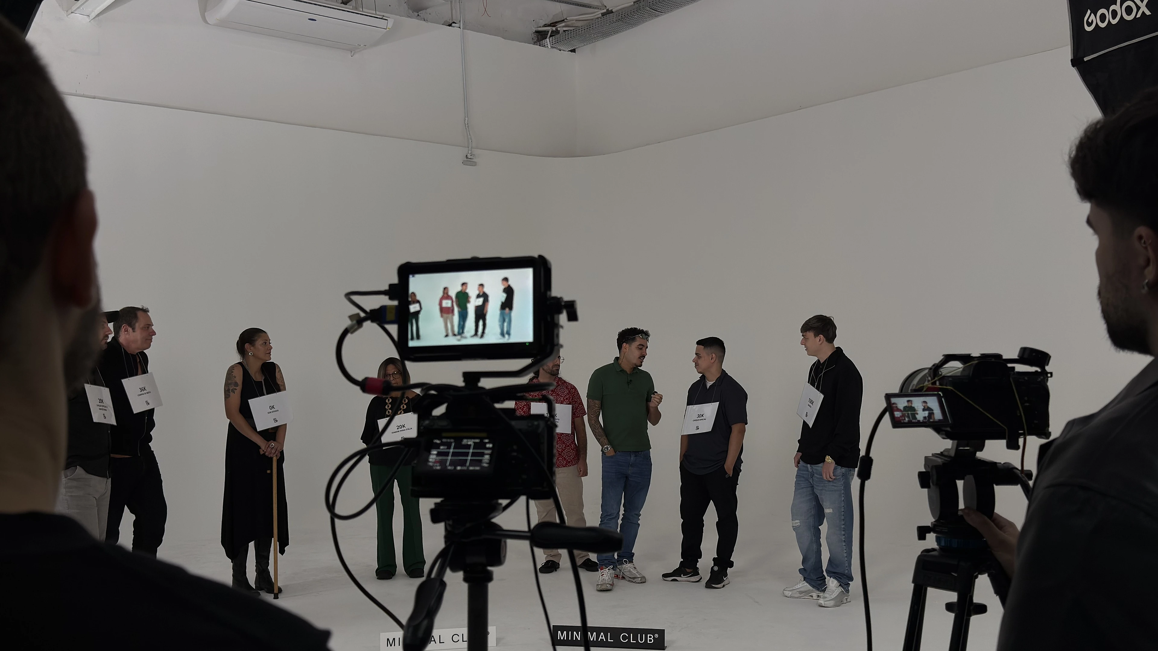 Câmeras em tripés e o monitor de uma delas, com os participantes em fila no estúdio branco ao fundo.