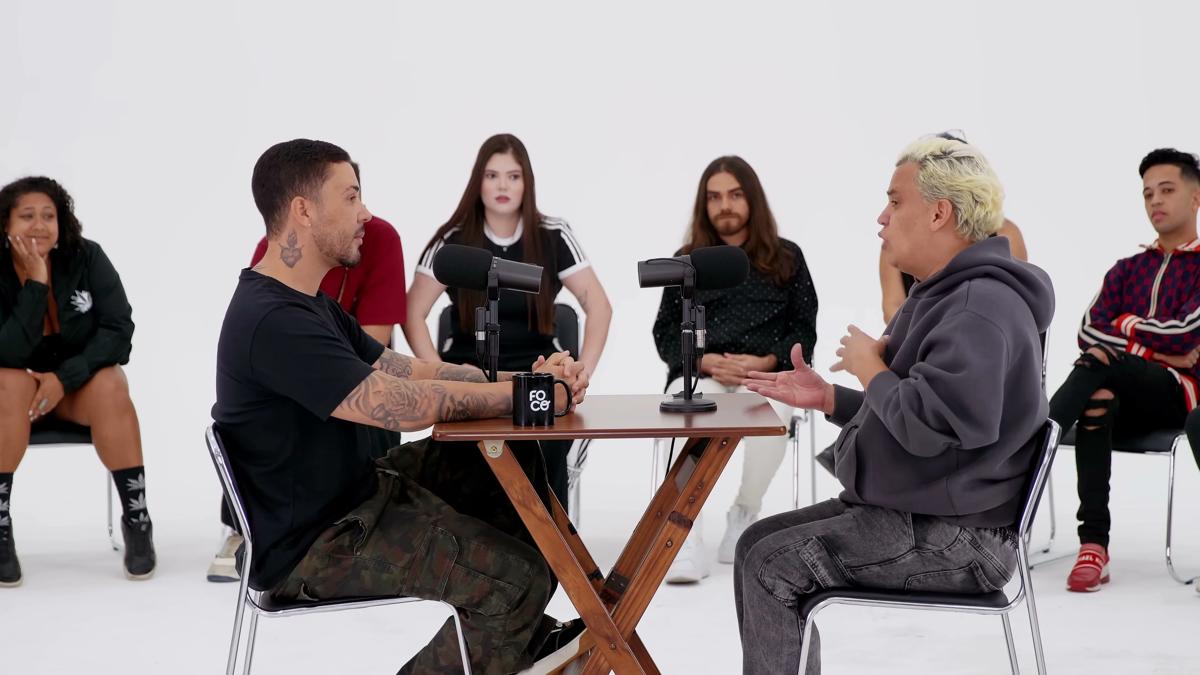 Dois homens conversam frente a frente na mesa de madeira do 1 vs 30, com microfones, e a roda de participantes sentada em volta, no estúdio branco.